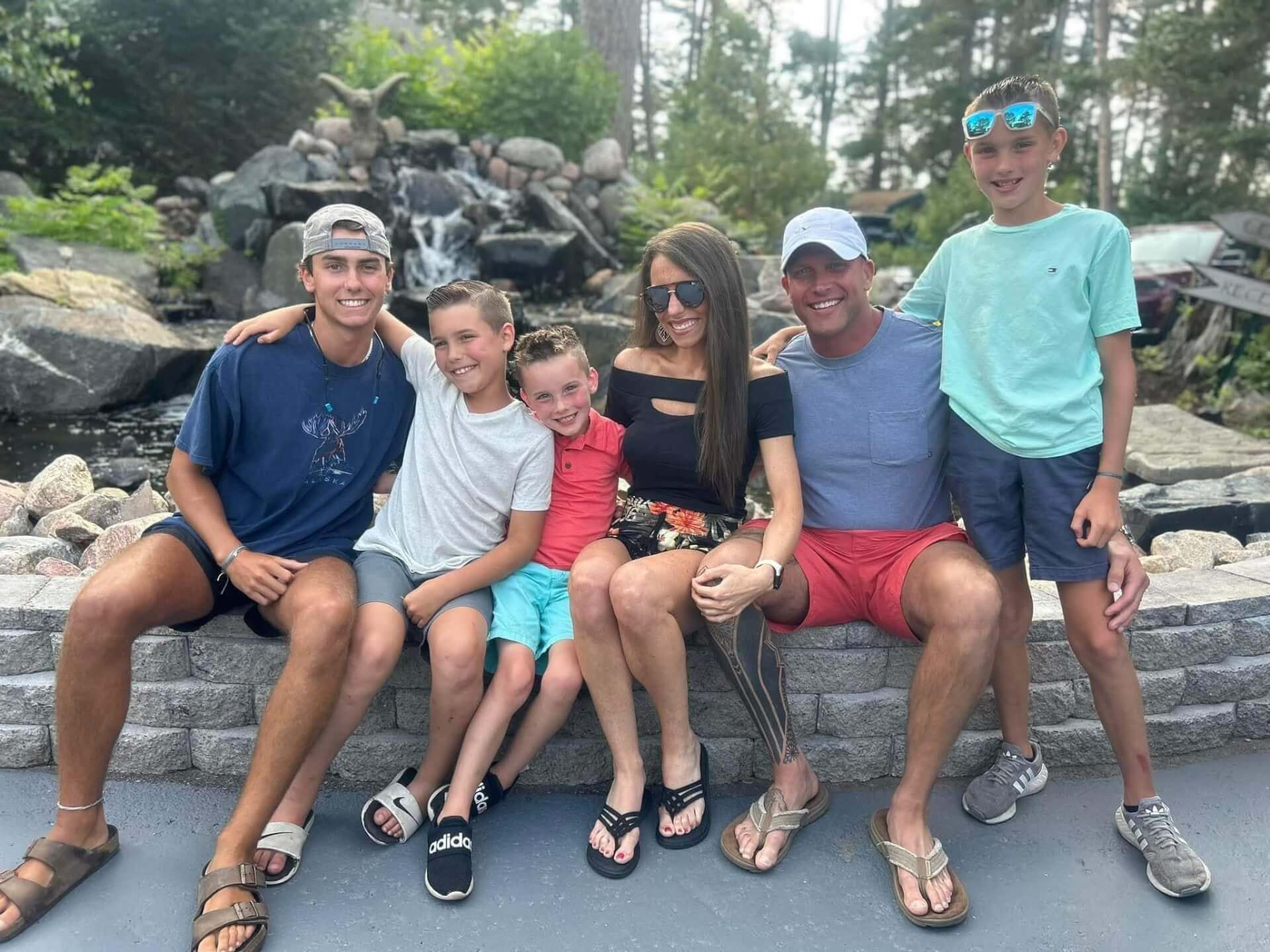 Multigenerational family of five smiling while seated on a stone ledge in front of a cascading waterfall and lush forest backdrop.