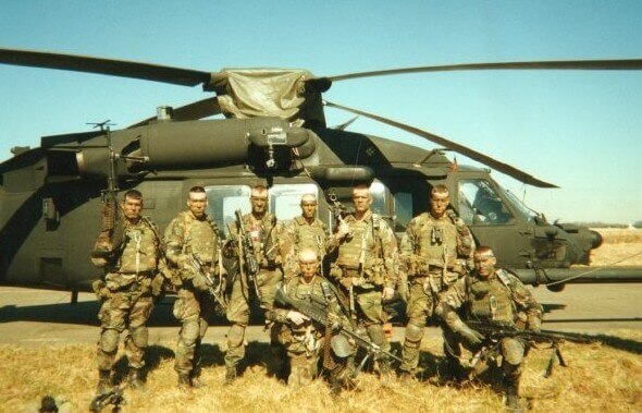 U.S. Army special operations soldiers in full combat gear standing in formation before a UH-60 Black Hawk helicopter on an airfield.