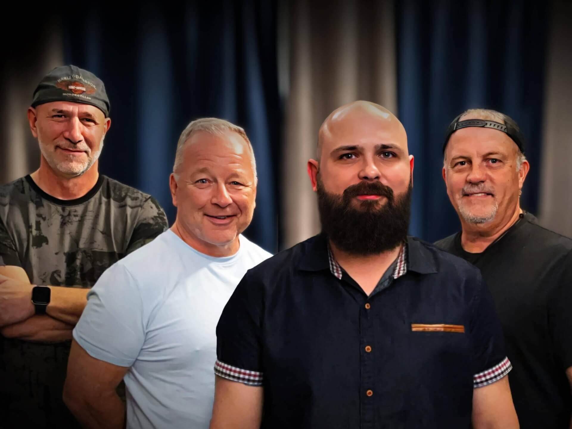 Group photo of four members of the rock band Doodz, featuring a bearded man in a dark shirt at the center, flanked by casually dressed bandmates, standing against a dark curtain backdrop.