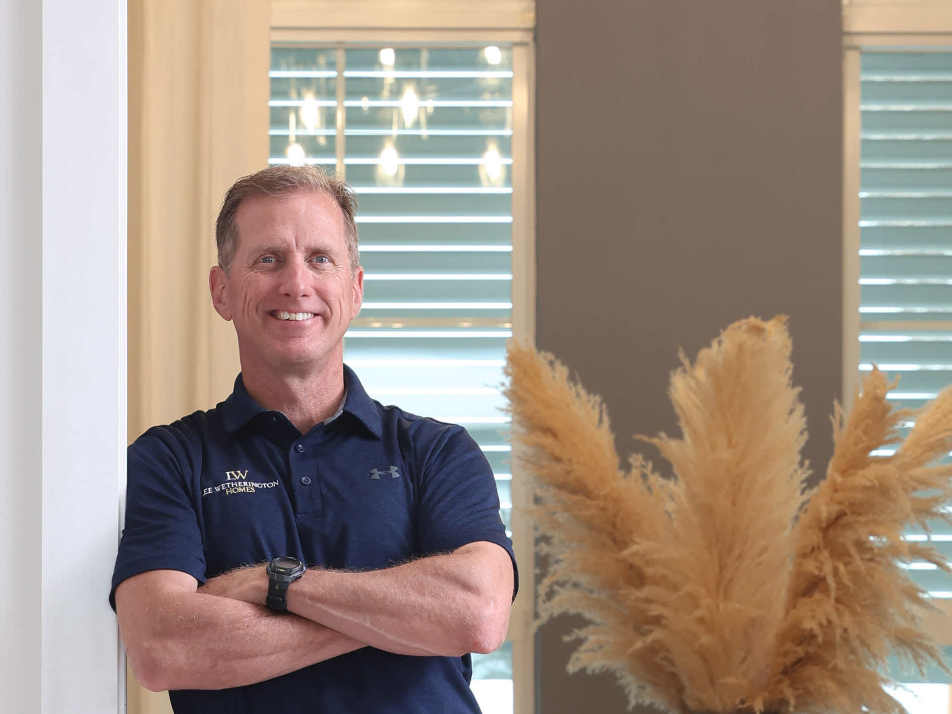 Smiling man in a dark blue Under Armour shirt with "Weatherington Homes" logo, arms crossed, in front of a wall of light-colored wood paneling.