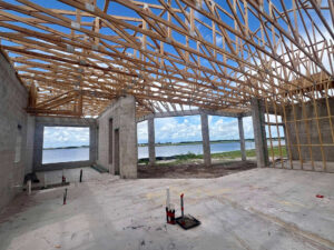 Interior view of a building under construction with a wooden roof frame and concrete block walls. A body of water is visible through the windows.