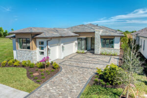Modern single-story home with a dark gray roof and a light gray stucco facade. A brick walkway leads to the front door, flanked by landscaping and a light fixture.