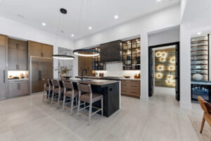 Modern kitchen with a large gray island, wooden bar stools, and dark wood cabinetry. A wine storage cabinet is visible in the background.
