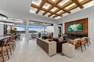 Modern living room with a light gray stone coffee table, leather chairs, and a large television on a dark wood wall. A dining area with bar stools is visible in the background.