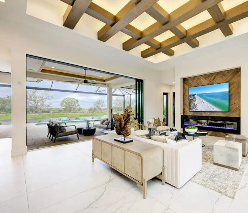 Spacious living room inside a luxury home in Florida with coffered ceiling, large TV wall, modern furniture, and sliding doors opening to an outdoor patio.