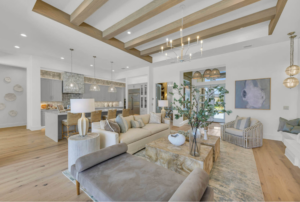 Bright open-concept luxury custom home living room with exposed wood ceiling beams, modern lighting, and elegant neutral furnishings flowing into a gourmet kitchen.