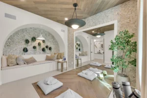 Luxury home wellness room with a built-in cushioned bench, pebble stone walls, wood ceiling, pendant lighting, floor cushions, indoor plants, and a large wall mirror.