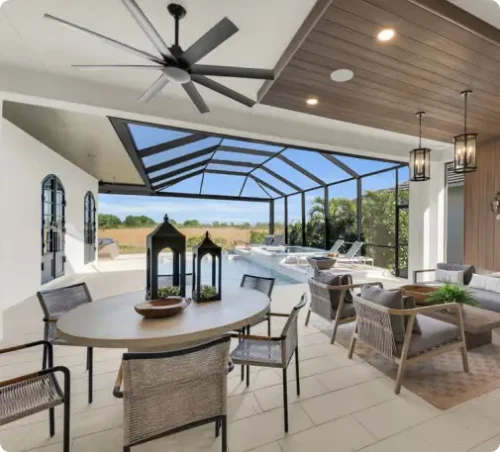 Outdoor patio and pool area of a luxury home with covered seating, dining table, ceiling fan, and screened enclosure overlooking the landscape.